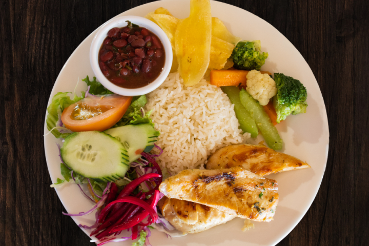 Plate with grilled chicken, rice, beans, salad, and vegetables on dark wood table.