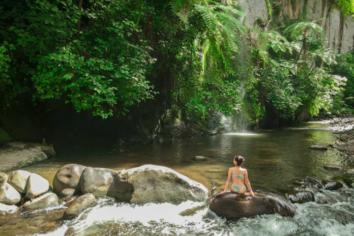 Person in swimwear sitting on a rock by a lush forest river with waterfall.
