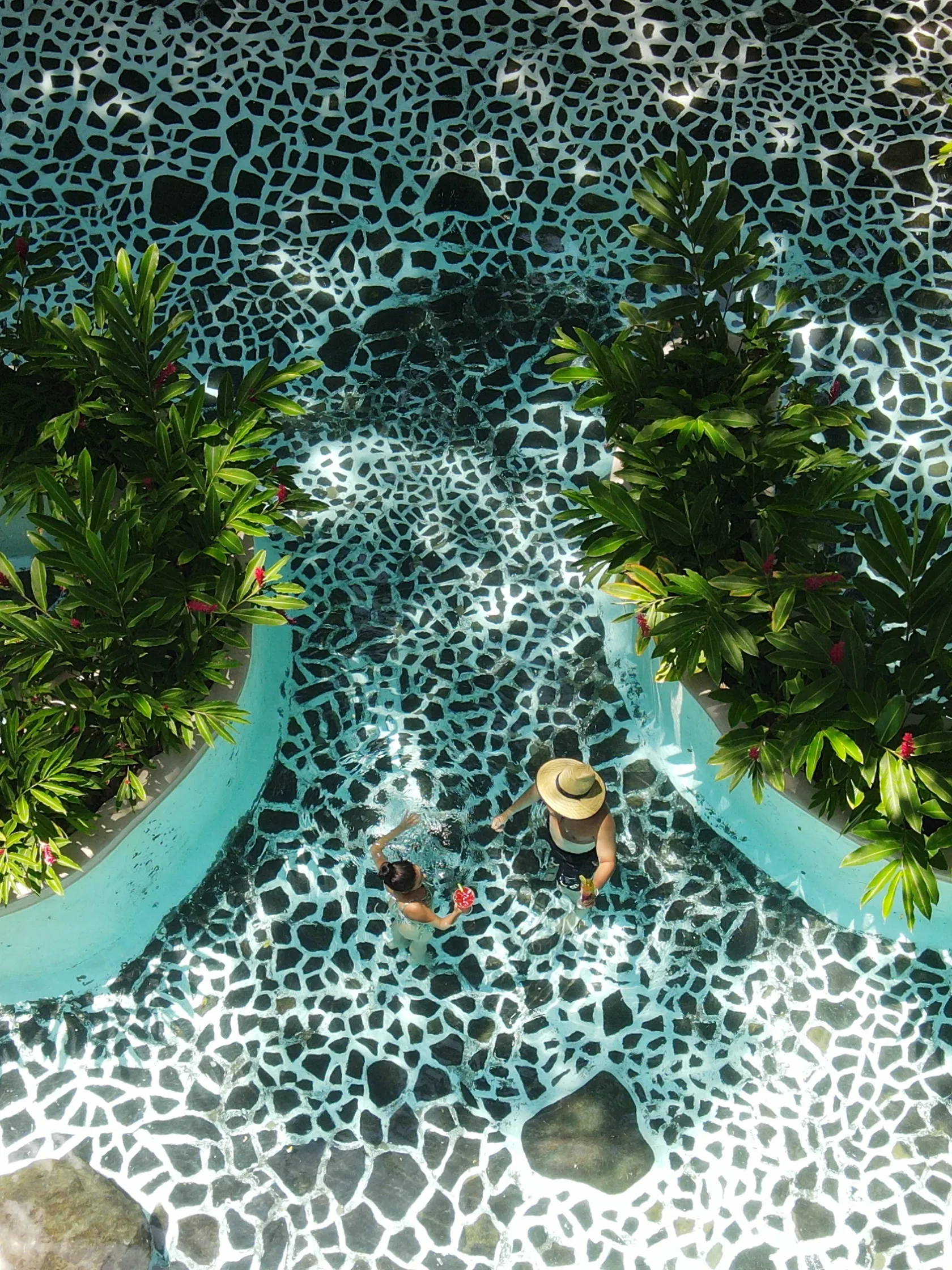Person in hat walking in pool with mosaic pattern and lush plant-lined path.