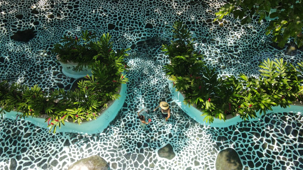 Person in hat walking in pool with mosaic pattern and lush plant-lined path.