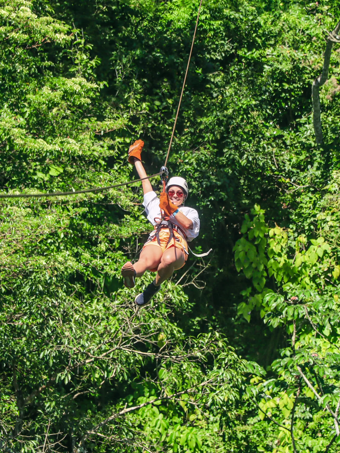 Person zip-lining through lush green trees, wearing helmet and gloves, smiling at camera.