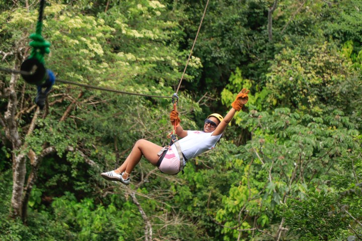 Person zip-lining through a forest, wearing helmet and gloves.