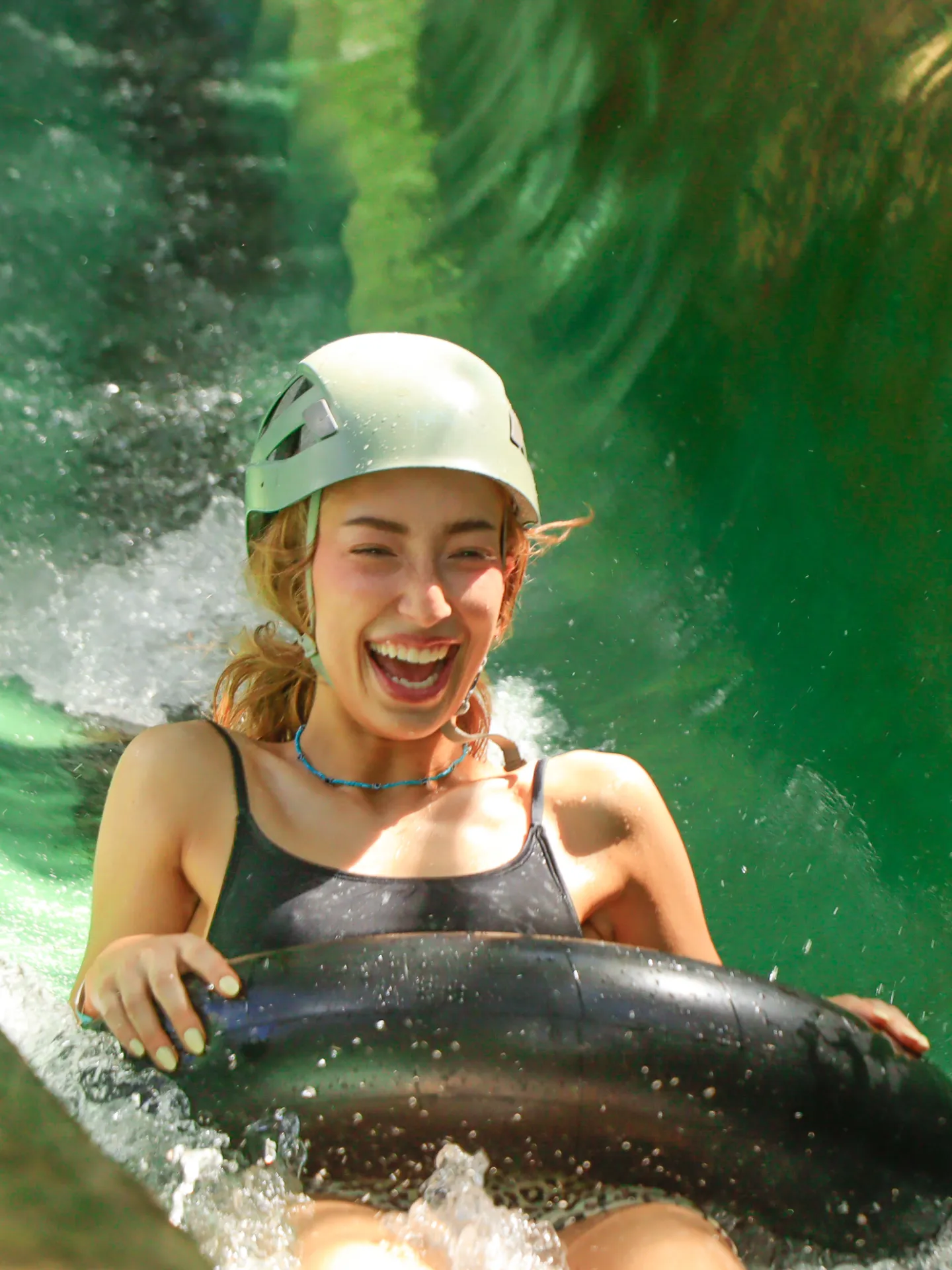 Person wearing helmet joyfully rides an inflatable tube down a green waterslide.