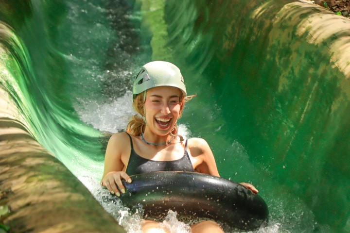 Person wearing helmet joyfully rides an inflatable tube down a green waterslide.