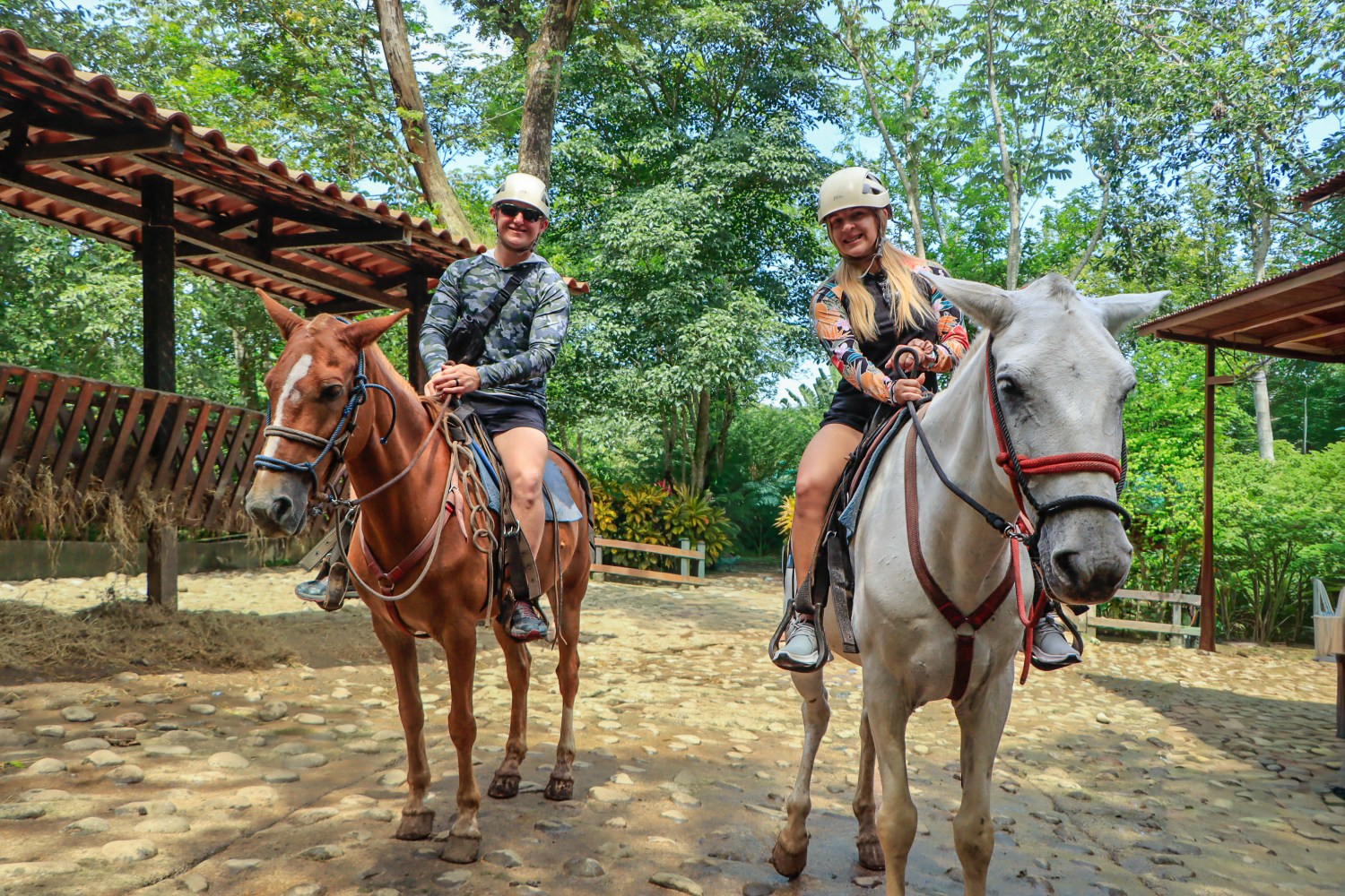 Two people riding horses under a canopy in a wooded area.