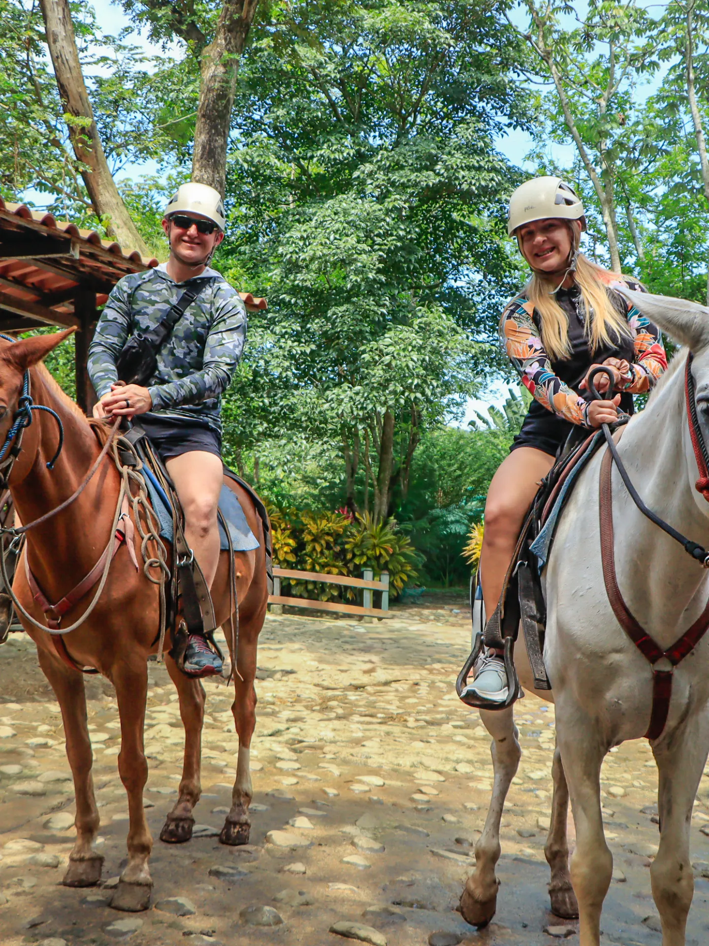 Two people riding horses under a canopy in a wooded area.