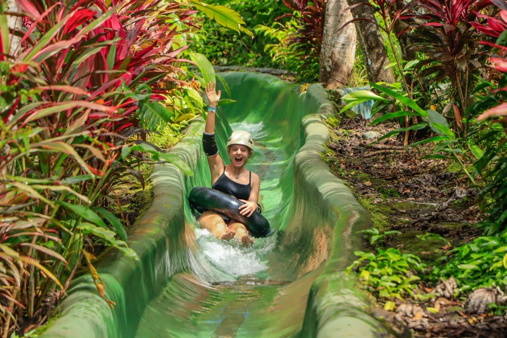 Person in black top sliding down a water slide surrounded by greenery, raising a hand in excitement.