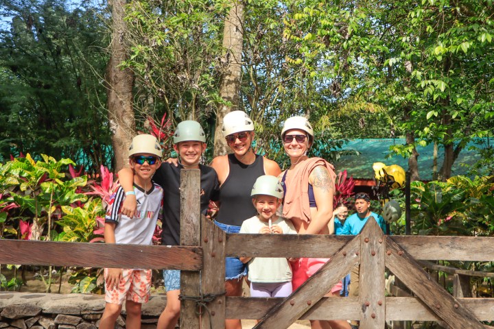 Group of five people wearing helmets and smiling outdoors, surrounded by trees and plants.