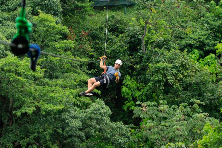 Person ziplining through dense green forest, wearing a helmet and gloves.