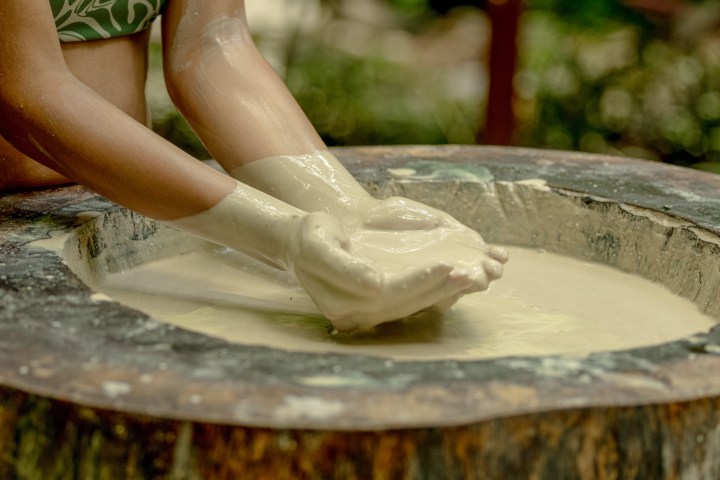 Hands mixing beige liquid in a large wooden bowl outdoors.