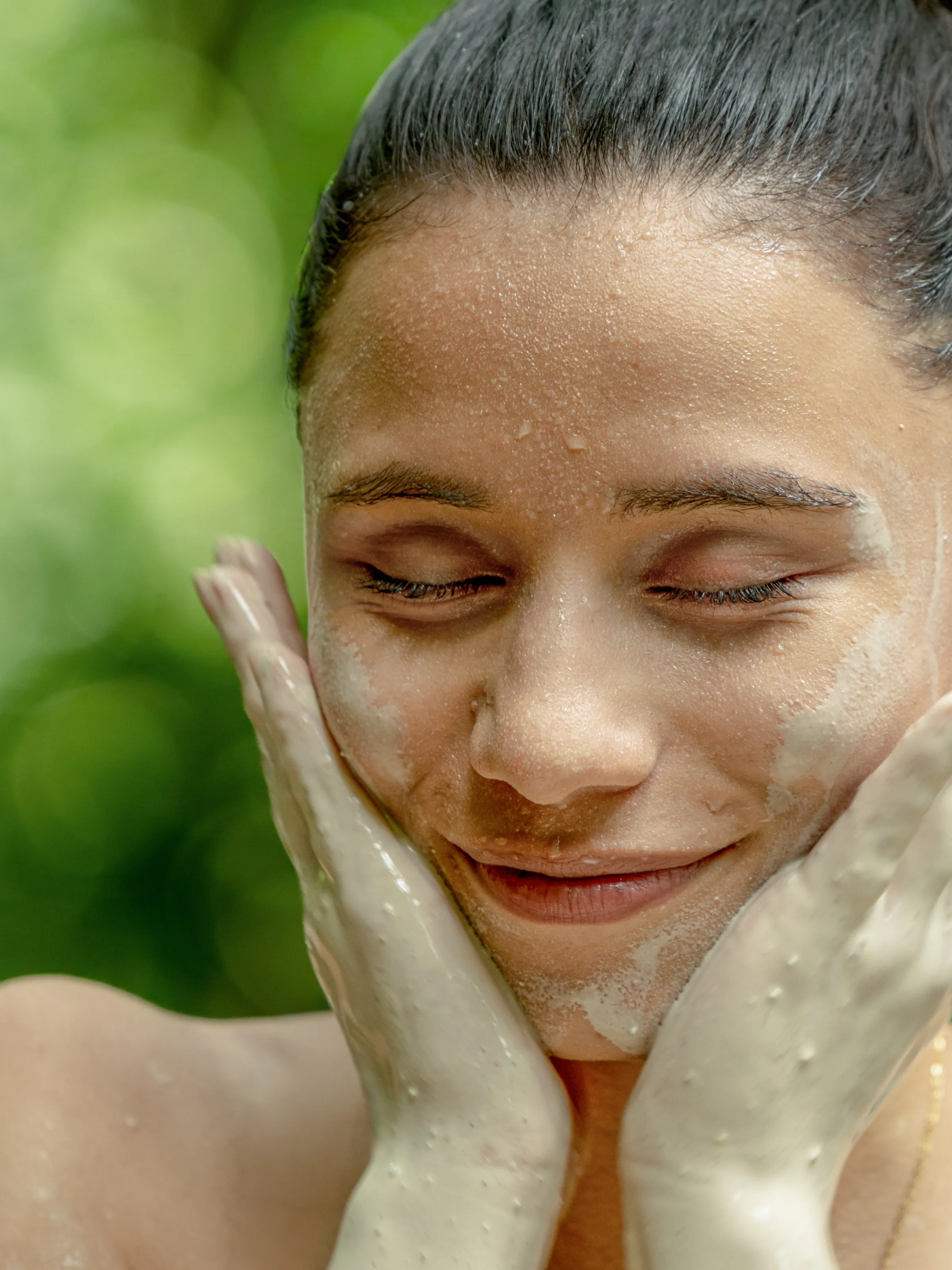 Woman with a clay mask on her face, smiling with eyes closed and hands on cheeks.