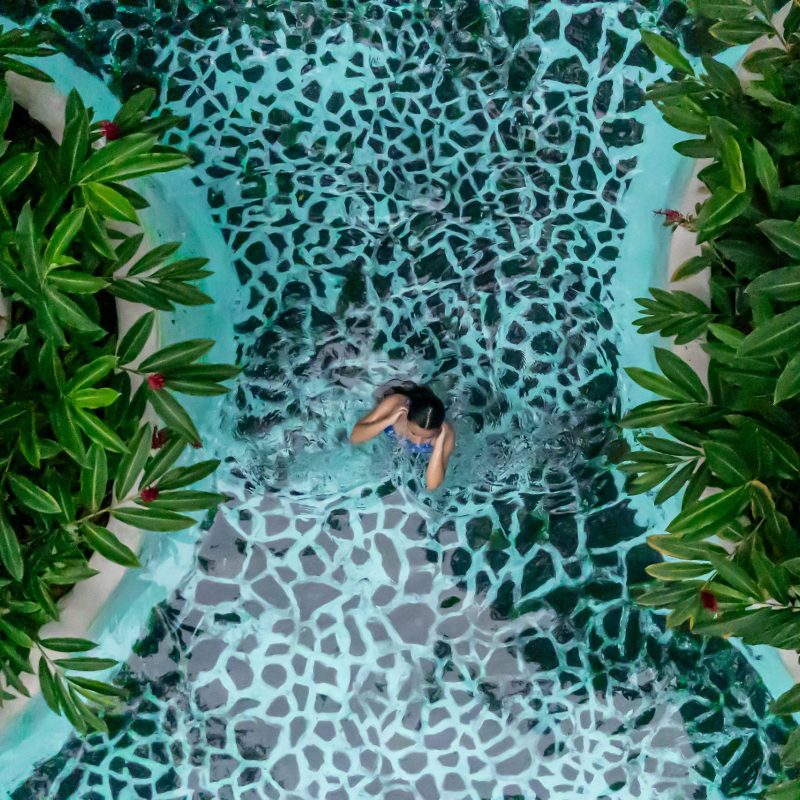Person swimming in a pool with a patterned floor, surrounded by lush greenery.
