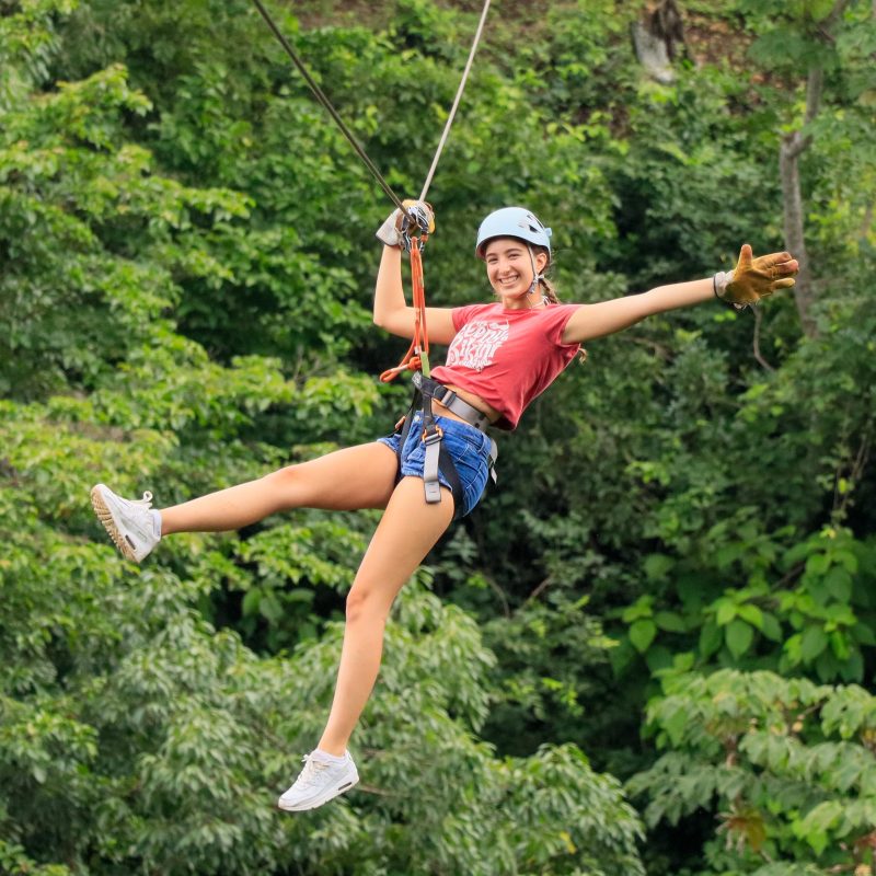 Person ziplining through lush green forest, wearing helmet and gloves.