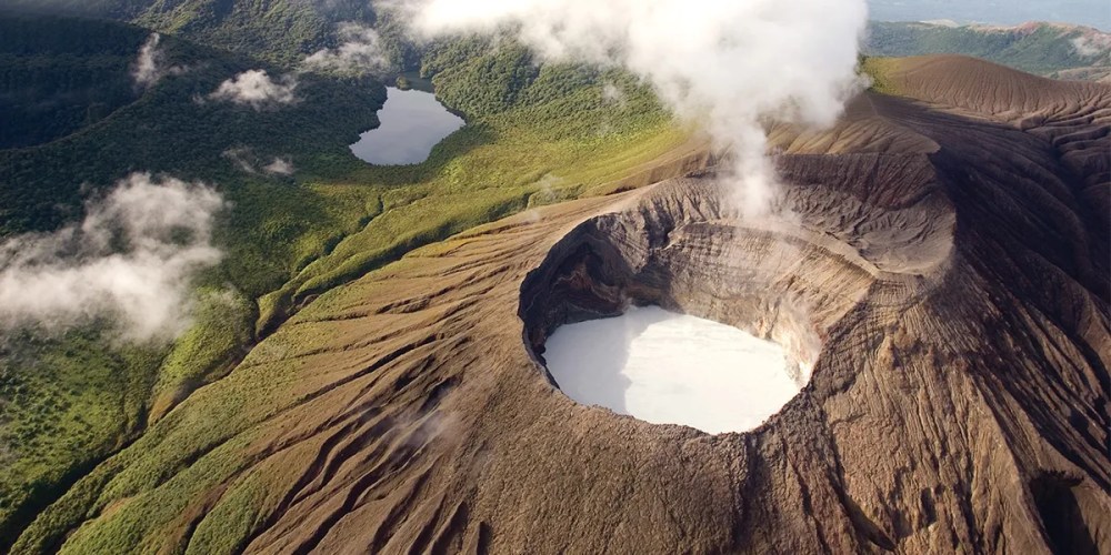 Aerial view of a smoking volcanic crater with a small lake surrounded by lush greenery and mountains.