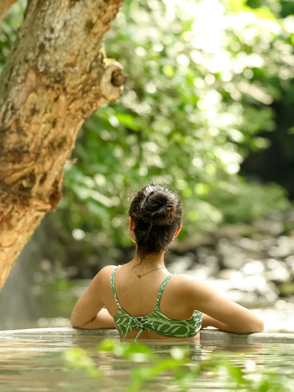 Person in a green swimsuit relaxes in a hot spring surrounded by lush greenery.