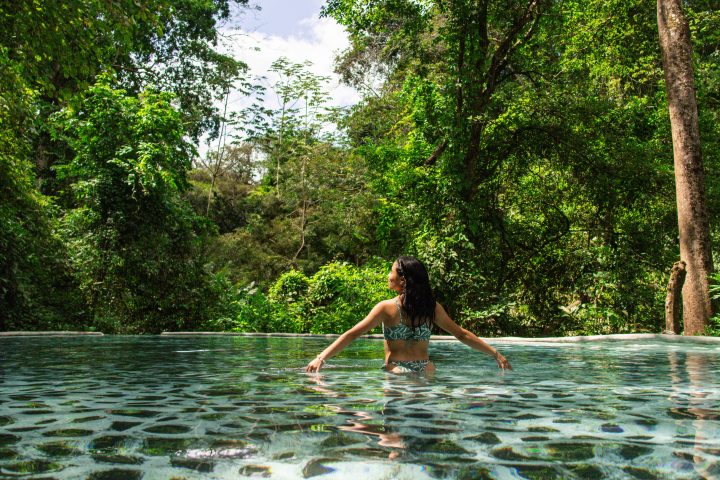 Person in swimsuit standing in a pool surrounded by lush green forest.