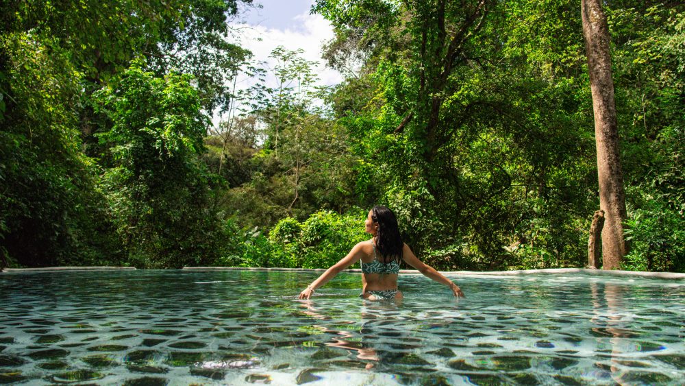 Person in swimsuit standing in a pool surrounded by lush green forest.