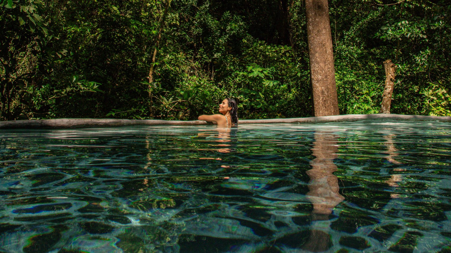 Person swimming in a clear pool surrounded by lush green trees.