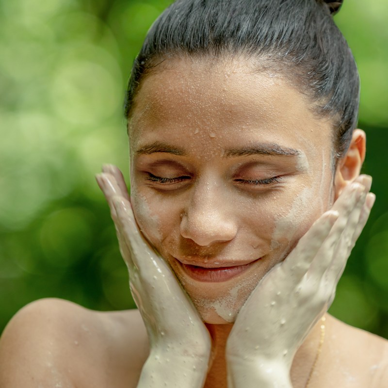 Woman with clay on face smiling with eyes closed, surrounded by greenery.